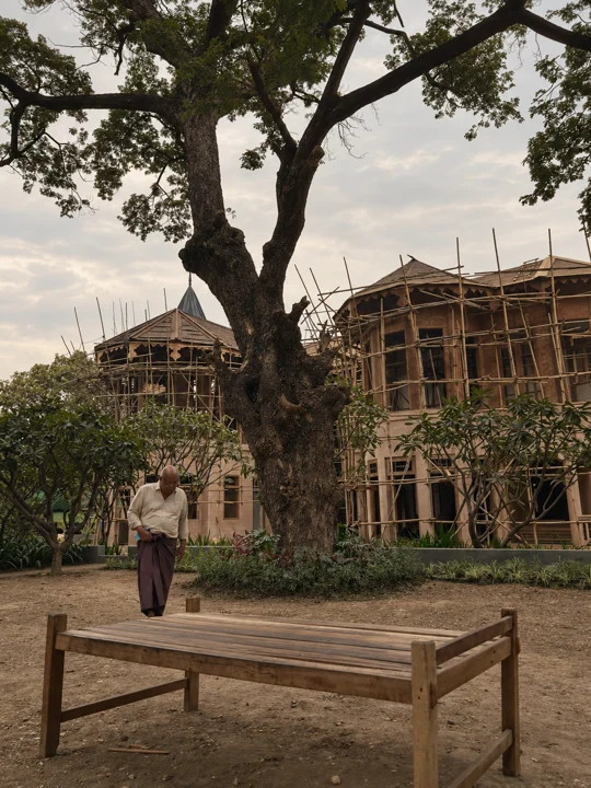 U Kyaw Sein walking beneath a mature tree in front of a scaffolded house under construction